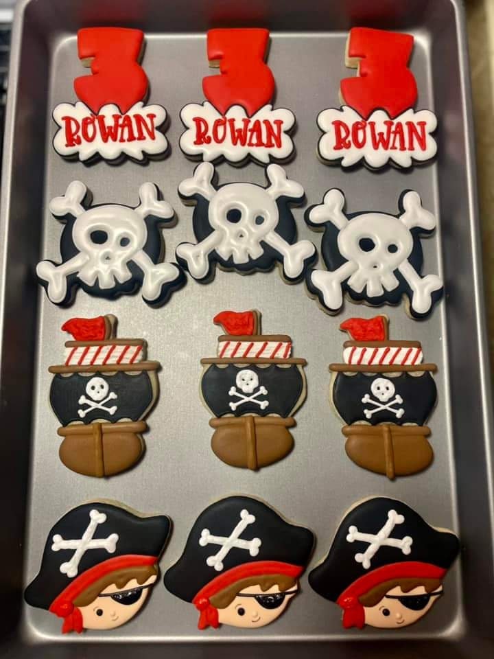 May include: A tray of nine decorated sugar cookies. Three cookies are red and white with the number '3' and the word 'ROWAN' on them. Three cookies are black, white, and red with a skull and crossbones design. Three cookies are black, white, and red with a pirate ship design. The cookies are arranged in three rows of three.