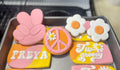 May include: A collection of six decorated cookies with a 1970s theme. The cookies are decorated with pink and yellow frosting and feature designs such as a peace sign, a flower, and the words 'Just groovy' and 'FREYA'.