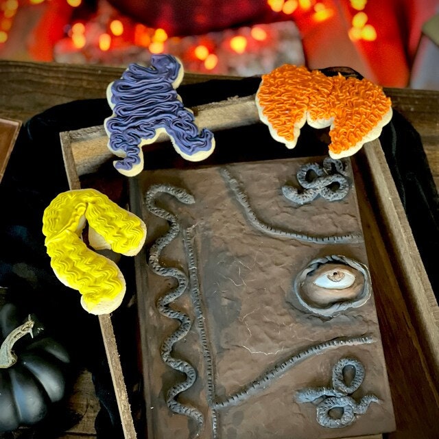 May include: Three Halloween cookies decorated with frosting. The cookies are shaped like a book, a snake, and a hand. The book cookie is brown with a black eye and snakes on the cover. The snake cookie is yellow and the hand cookie is purple.